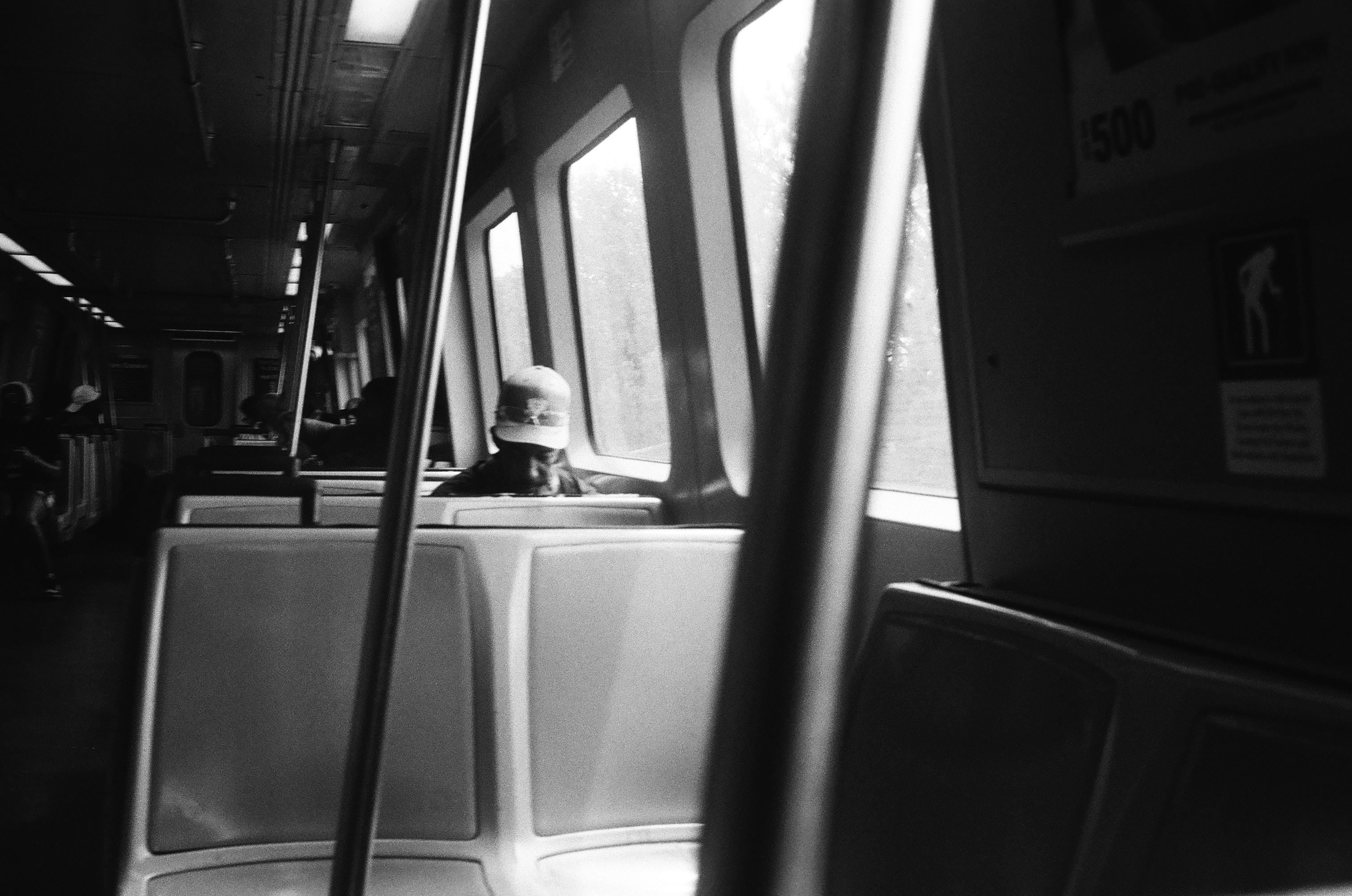 B&W photo of a person seated on a subway train, a ballcap on their head. The sun shines in from the bank of windows besides them, marked against the dim interior of the railcar. Two metal railings strike down from the ceiling near the viewer.